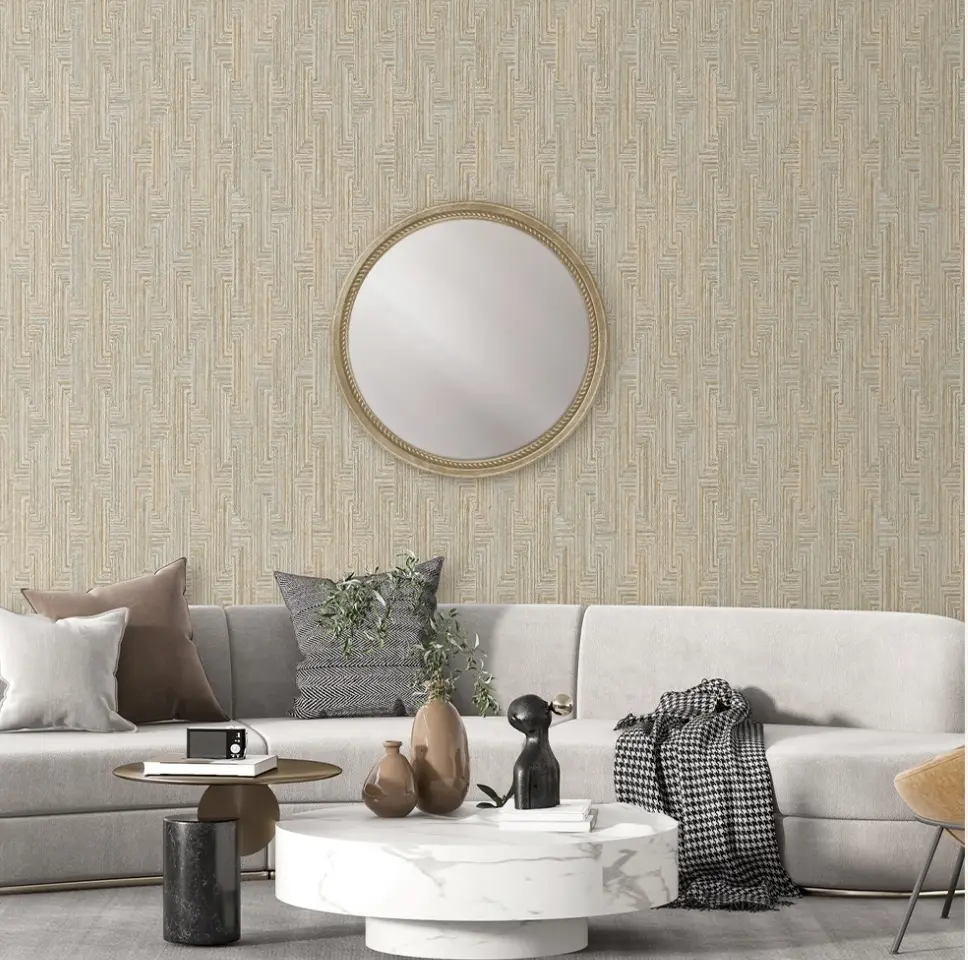 Un sofa curvo gris clarito decorado con cojines en las tonalidades marrón y gris. Enfrente encontramos una mesilla de mármol blanca. el fondo encontramos una pared de papel pintado con formas geométricas beige y un espejo redondo dorado en el centro.